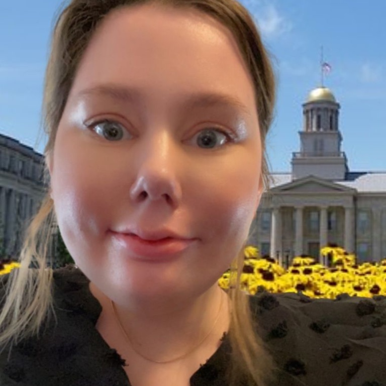 White woman with shoulder-length blonde hair wearing a black blouse with ruffled neck. Behind her is the University of Iowa Old Capitol building.